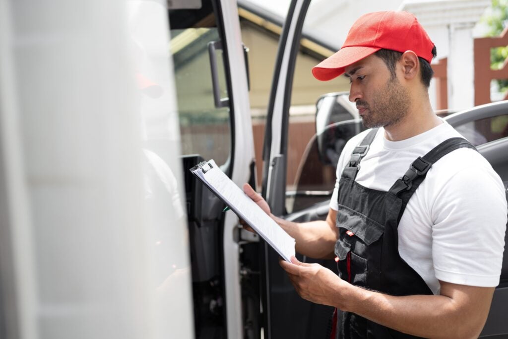 A transport worker in a red cap and overalls checking a clipboard next to a vehicle