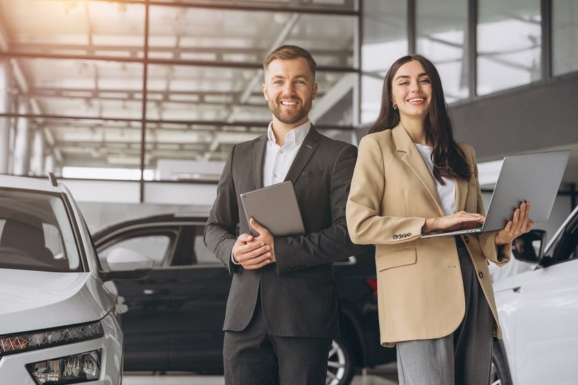 A couple holding a tablet and laptop, standing by their cars, preparing to sell their personal vehicles online.